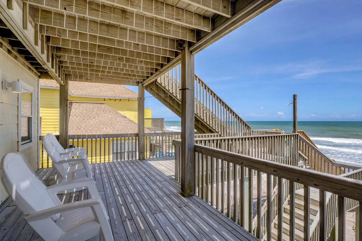 A spacious wooden deck is furnished with white Adirondack chairs facing the ocean. A staircase leads directly down to the sand, and the vast sea stretches out to the horizon, bathed in soft, golden evening light. The setting is tranquil and inviting, perfect for relaxing or socializing while taking in panoramic ocean views.