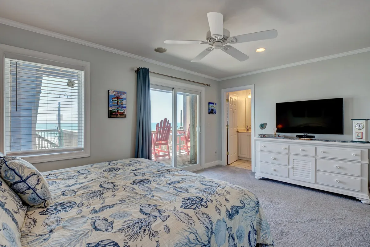 A tranquil master bedroom with blue seashell-patterned bedding, a large sliding glass door that opens to outdoor seating, and a spacious dresser with a television. The room is bathed in natural light and offers views of the beach just beyond the deck.