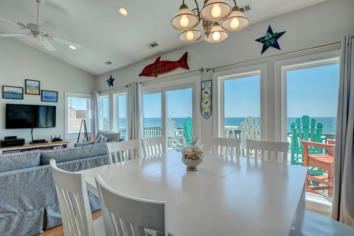 A cheerful dining area with a large table surrounded by chairs, set in front of picture windows offering panoramic views of the ocean. Comfortable seating and beach-themed décor create a welcoming space for shared meals or game nights.