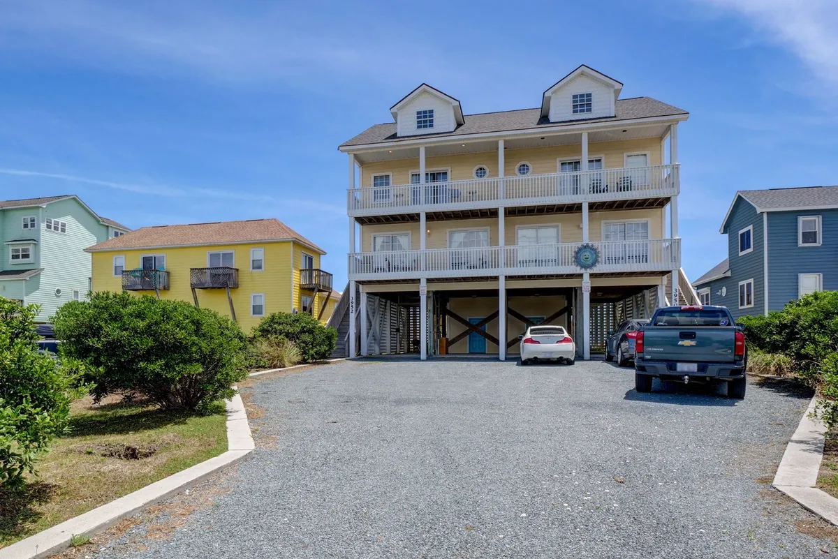 A cheerful exterior view of a vibrant three-story yellow house with balconies featuring railings, colorful neighboring homes nearby, and vehicles in the driveway. The scene suggests a welcoming and spacious environment ideal for family gatherings and group activities.