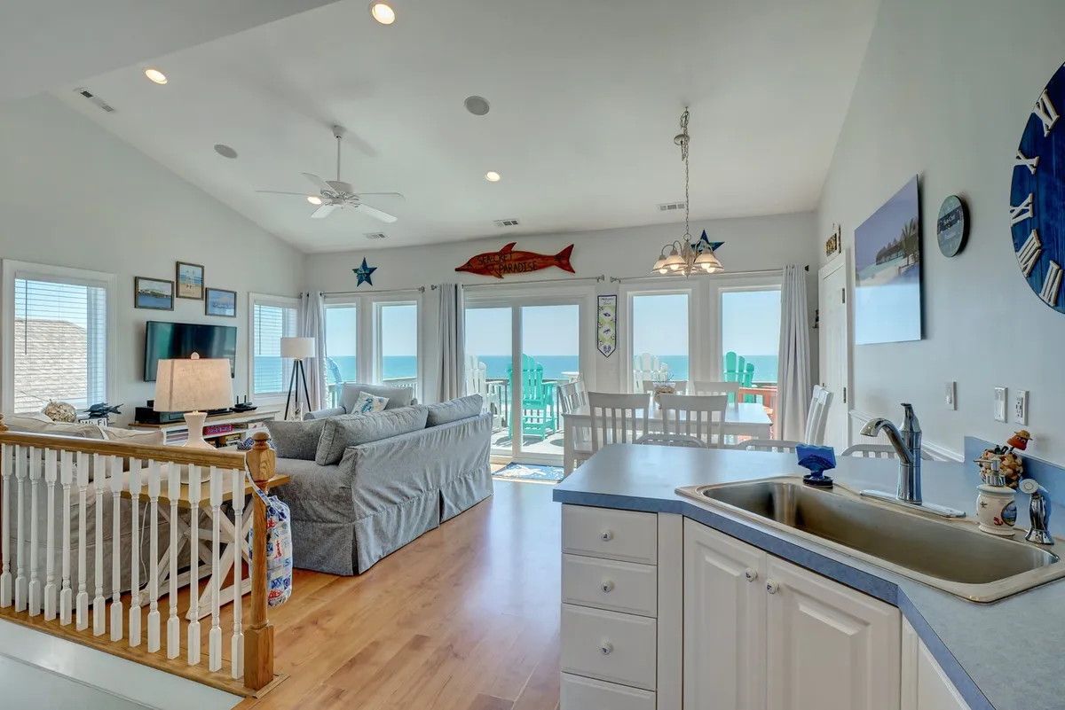 A wide-angle view of a spacious living area with a comfortable sofa, a dining table set in front of large windows overlooking the ocean, decorative fish accents on the wall, and an open kitchen counter visible to the side. The space feels bright, airy, and ideal for group gatherings.