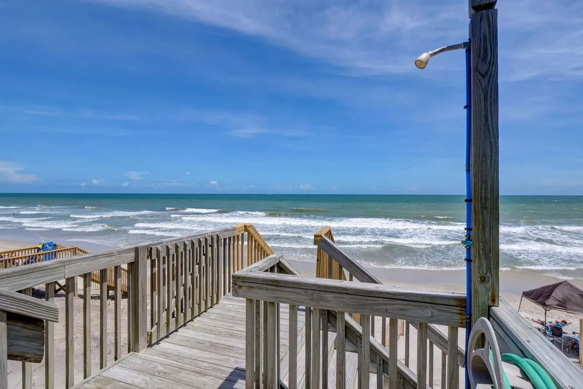 A broad boardwalk leads through gentle dunes to the sandy beach, with the ocean and distant umbrellas visible beyond. The scene is serene and lively, capturing the essence of direct beach access and the potential for family activities like sandcastle building or sunset picnics.