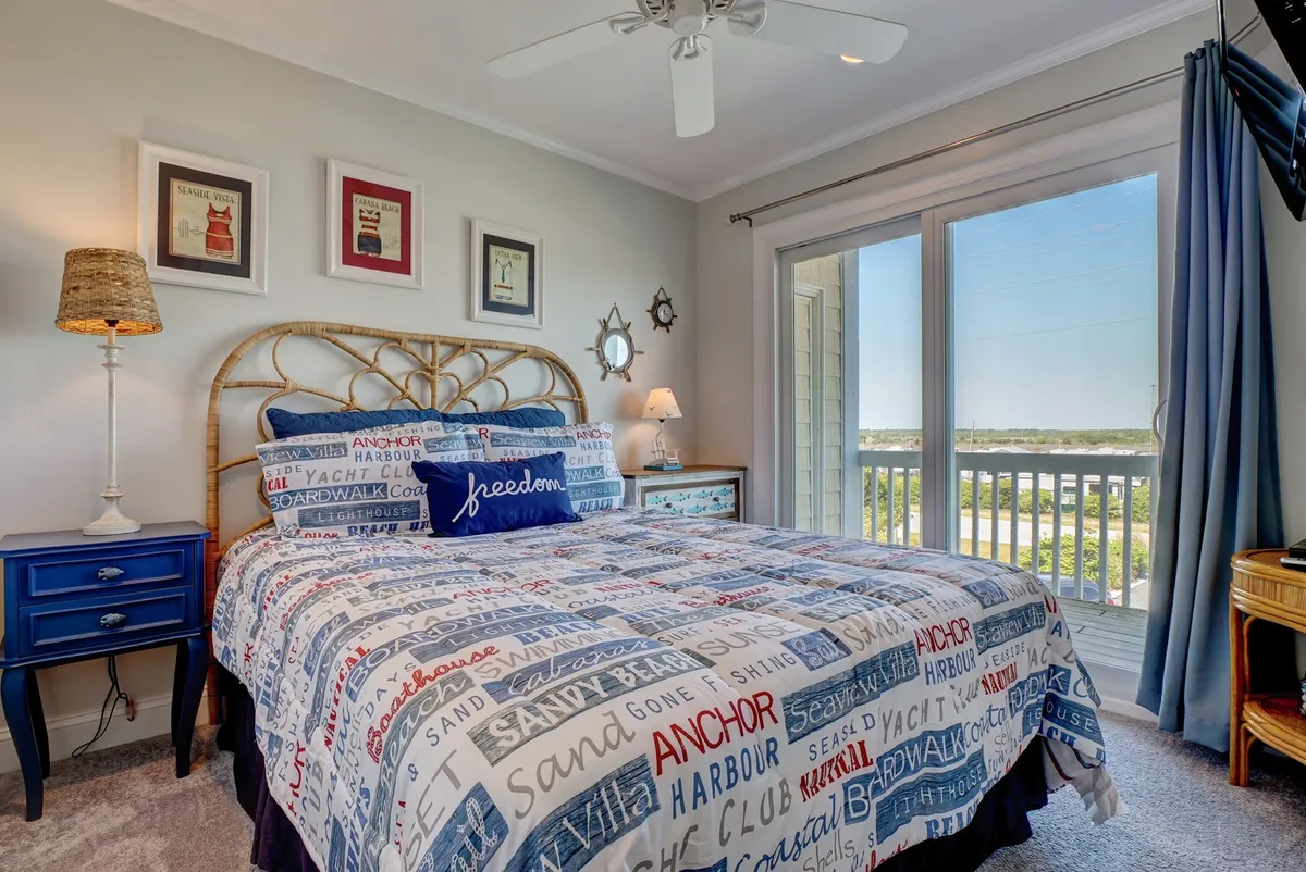 A serene bedroom featuring a bed with a patterned duvet, a calming blue nightstand, and a large window that lets in natural light. The window suggests an upper-level location, and there is a hint of a view that could correspond to a private balcony.