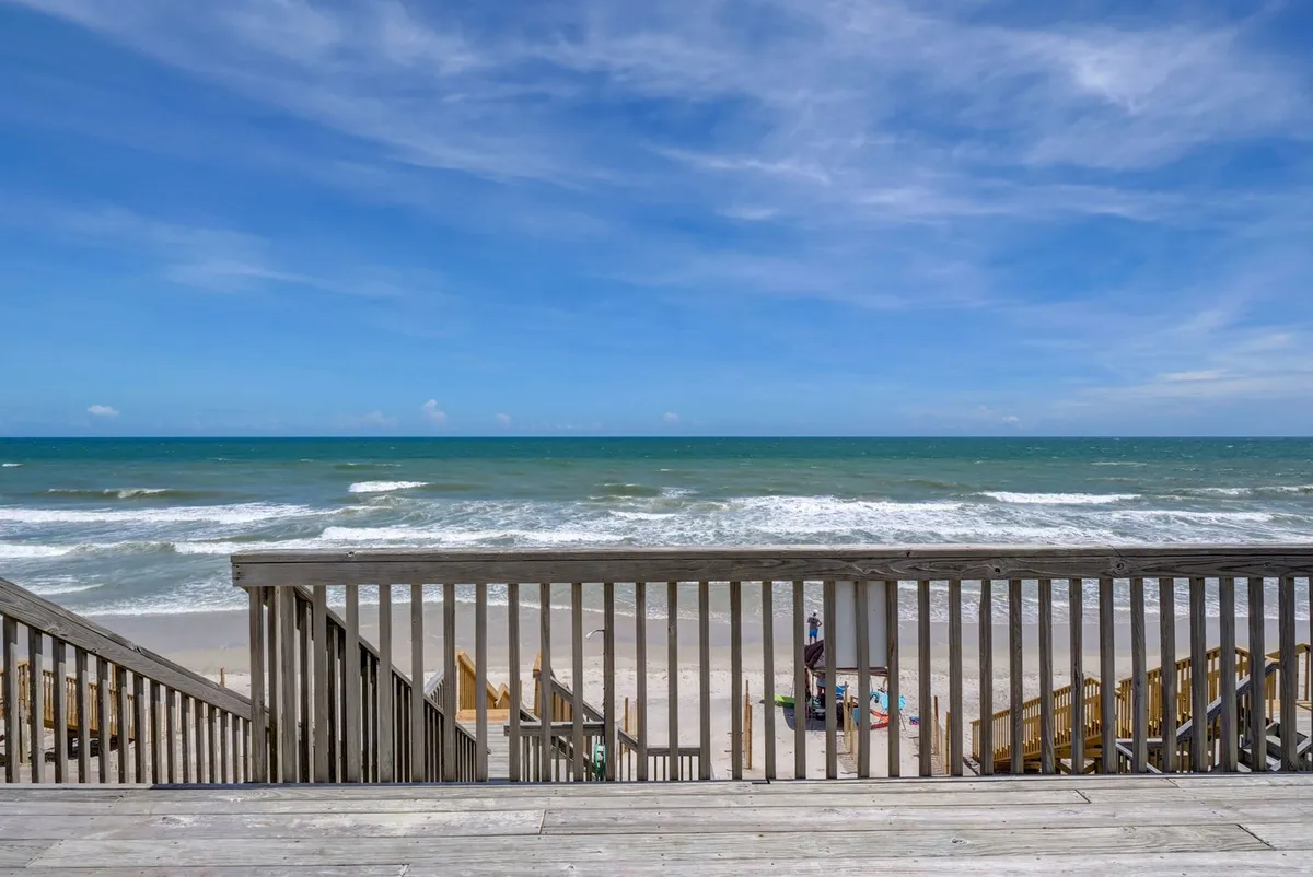 A tranquil scene of a wooden deck overlooking the beach, complete with beach chairs set up for sunbathing or relaxing. The view extends across rolling ocean waves under a cloudy sky, emphasizing the home's prime beachfront location and the inviting outdoor lounging setup.