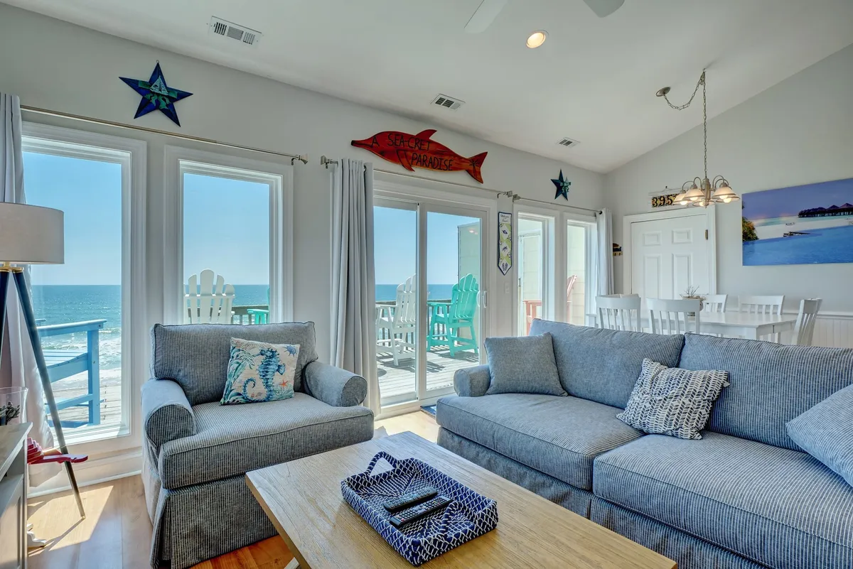 A bright, cheerful living room with large windows offering unobstructed ocean views, comfortable seating, and playful coastal decor (fish and starfish). The space invites relaxation while soaking in sunrise views or watching the waves.