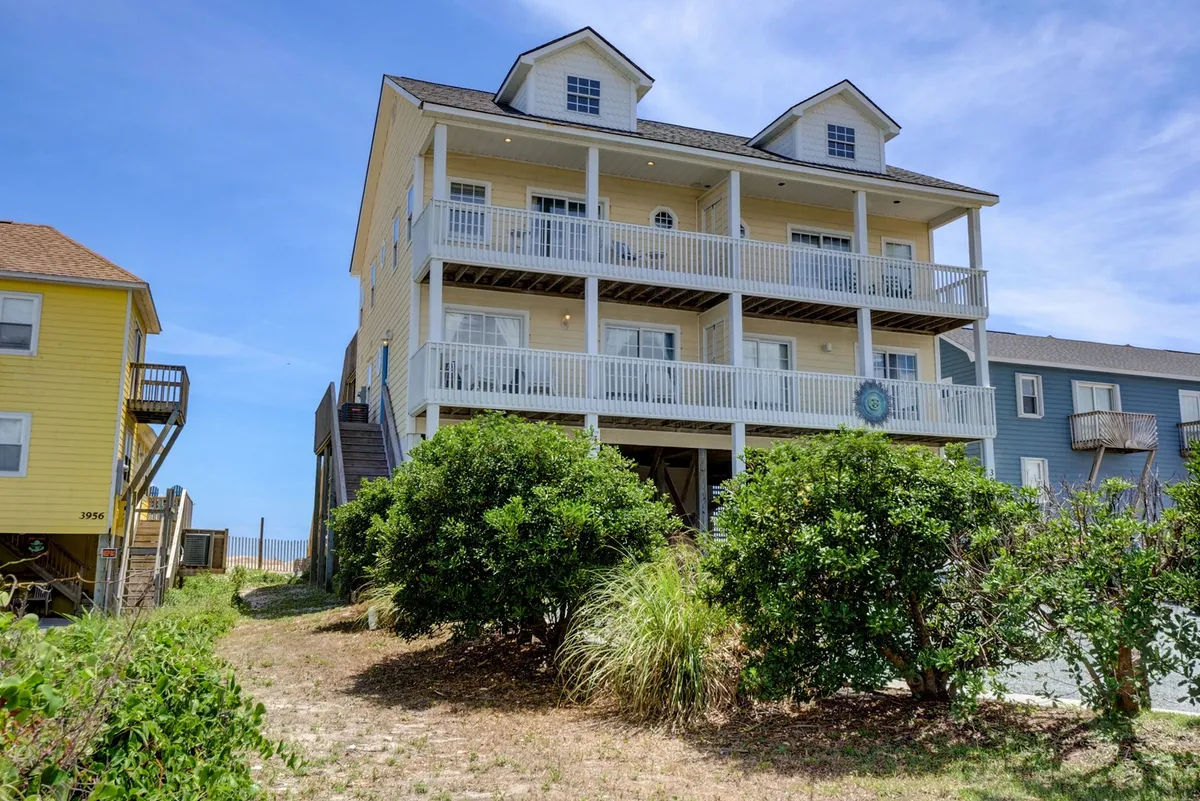 An exterior view of a cheerful three-story house bordered by lush greenery, with balconies overlooking the landscape and adjacent houses visible nearby. The inviting facade and spacious decks hint at relaxation and a welcoming space for families to gather and enjoy the outdoors.