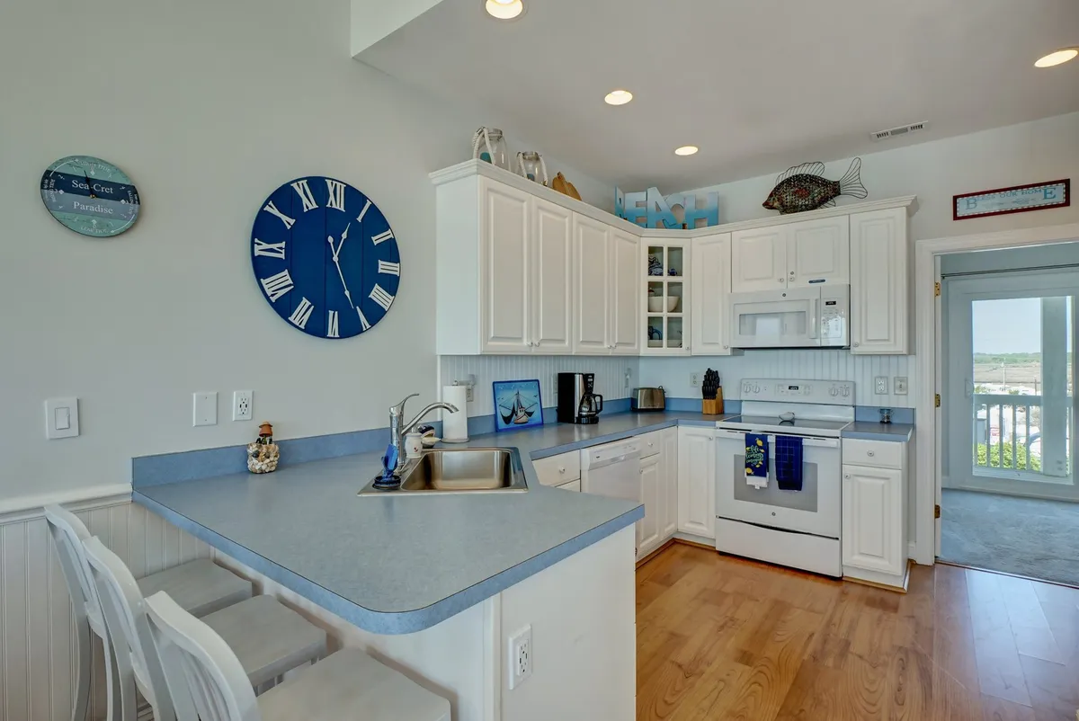 A modern kitchen with white cabinetry, a kitchen island, a blue clock on the wall, and an open door leading to a balcony or deck. The space is filled with natural light, encouraging social cooking and easy movement between interior and exterior.