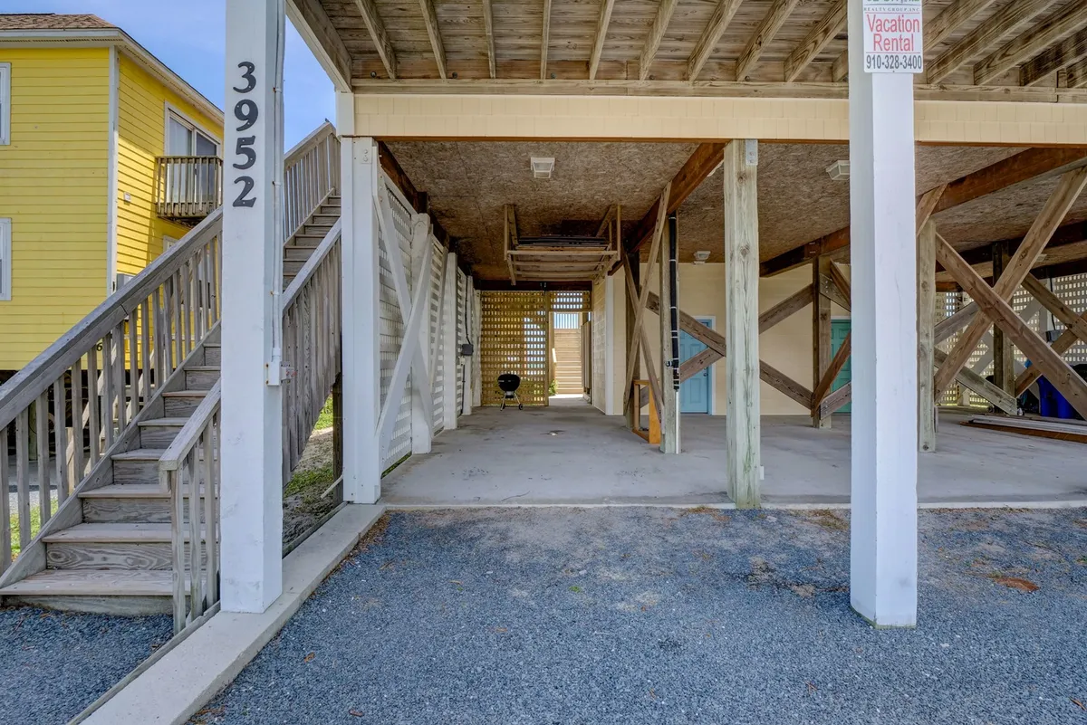 This image captures the exterior of the property, featuring a staircase leading up to the house, a vacation rental sign, open space beneath the house, and a gravel driveway. The relaxed vibe suggests a place to gather, park after beach excursions, or enjoy sunset barbecues.