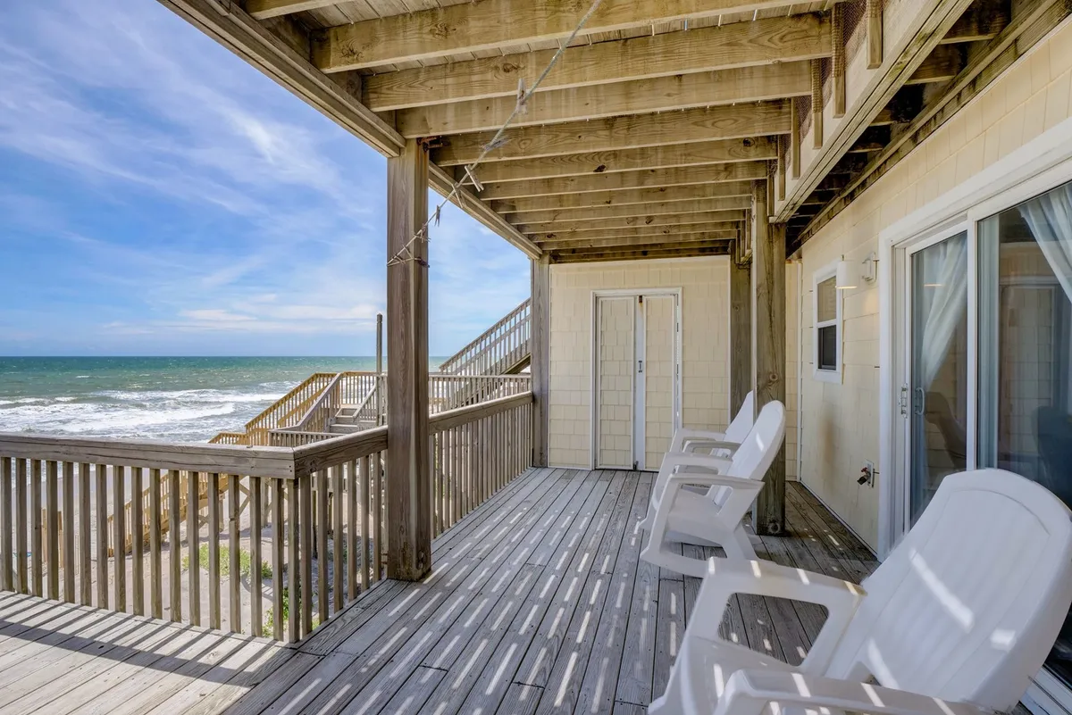 A tranquil deck scene featuring wooden Adirondack chairs facing out to a sweeping beach view, with a wooden deck rail framing the expansive ocean and sky. The setting is perfect for morning coffee, reading, or simply soaking in the sights and sounds of the coast.