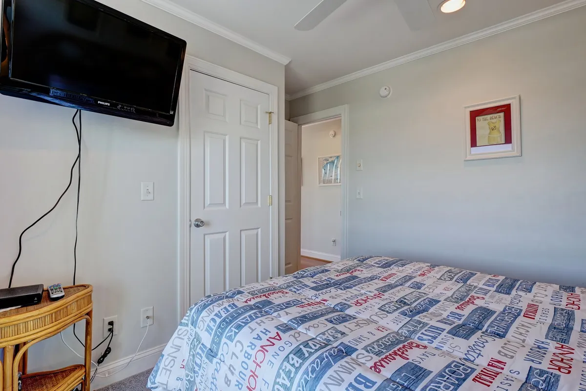 A cozy guest bedroom featuring a queen bed adorned with patterned bedding. The room includes a wall-mounted TV, a door leading to the bathroom, framed artwork, and a woven side table, creating a tranquil and inviting space.