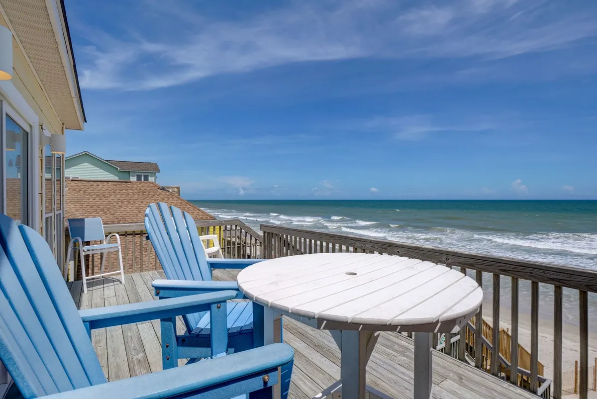 The image showcases a serene beachfront deck with inviting blue chairs and a white table, offering unobstructed views of the ocean. The scene radiates tranquility, with the promise of relaxing mornings sipping coffee, long conversations while soaking in the sea breeze, and breathtaking sunrises over the water. The setting is perfect for enjoying meals outdoors, unwinding with loved ones, and reconnecting with nature in a peaceful, coastal environment.